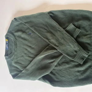 Grön stickad tröja Polo Ralph Lauren - Mörkgrön stickad tröja från Polo Ralph Lauren med rund halsringning och diskret broderad logga på bröstet. Klassisk modell med ribbade muddar vid ärmslut och nederkant. Perfekt för lager-på-lager under hösten och vintern.