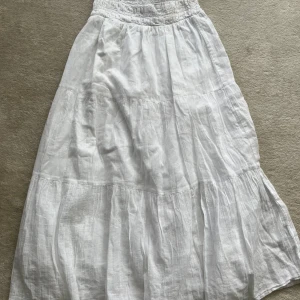 Vit långkjol från Gina Tricot - Luftig och flowy vit långkjol från Gina Tricot med smockad midja och flera volanger. Kjolen är tillverkad i ett tunt och svalt tyg, perfekt för varma dagar. Enkel att styla med crop top eller linne för en chill sommarlook.