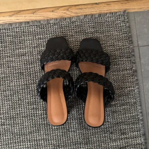 Svarta flätade sandaler med klack - Snygga svarta sandaler med två breda flätade remmar och fyrkantig tå. Skorna har en låg klack och öppen häl, vilket ger en trendig look. Tillverkade i syntetmaterial med en stilren design som passar perfekt till sommarens outfits. Pernilla Wahlgren kollektion. Storlek 41. Men skulle säga att de passar bra på en som har 39/40