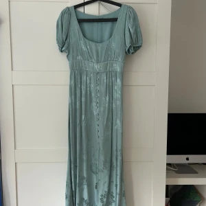 Ljusgrön långklänning från Zara - Superfin ljusgrön långklänning från Zara med puffärmar, blommigt mönster och knappar framtill. Klänningen har markerad midja, slits och dragkedja i sidan. Defekt på bild 5 