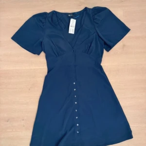 Mörkblå kortklänning med knappar - Supersnygg mörkblå kortklänning med v-ringning och korta puffärmar. Klänningen har knappar längs hela framsidan. Den är endast testad och har prislappen kvar.💕                       Den kostar 400kr ny