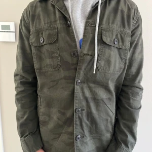 Grön camo overshirt med knappar - Snygg militärgrön overshirt i camomönster med två bröstfickor och knäppning framtill. Jackan är i bomull och har en avslappnad passform, perfekt att slänga över en hoodie. Långärmad och med klassisk krage. Funkar perfekt för sena sommarkvällar samt utgångar. Jackan är i 9/10 skick och säljs inte längre på hemsidor. 