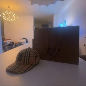 Burberry rutig keps med logga - Snygg keps från Burberry i klassiskt beige rutigt mönster med svarta, vita och röda ränder. Framtill finns en svart broderad Burberry-logga. Justerbar rem baktill med metallspänne. Perfekt accessoar för att lyfta din outfit.