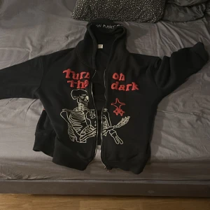 Svart hoodie Broken Planet Market - Svart hoodie från Broken Planet Market med dragkedja, huva och broderad text på luvan. Framsidan har ett stort skelett-tryck och röd text, samt röda stjärndetaljer både fram och bak. Oversized fit och riktigt streetwear-vibe.