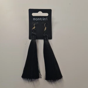 Svarta tofsörhängen från Montini - Långa svarta örhängen från Montini med trendiga tofsar i glansigt material. De har guldfärgade krokar och ger en cool statement-look. Perfekta för att lyfta vilken outfit som helst.