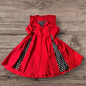 Röd retroklänning från iHot - Supersnygg röd klänning från iHot med svartvita prickiga detaljer i kjolen och vid halsen. Klänningen är ärmlös, har dekorativa svarta knappar framtill och en v-ringad krage. Materialet känns som bomull och modellen har en klassisk retrovibe.