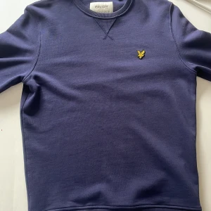 Marinblå sweatshirt från Lyle & Scott - Snygg marinblå sweatshirt från Lyle & Scott i ekologisk bomull. Klassisk rund halsringning, ribbade muddar och ikonisk gul logga på bröstet. Perfekt för dig som gillar stilren och enkel design med en sportig touch.