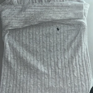 Grå kabelstickad tröja Polo Ralph Lauren - Snygg grå kabelstickad tröja från Polo Ralph Lauren med klassisk broderad logga på bröstet. Tröjan har ribbade muddar och är perfekt för lager-på-lager. Den stickade strukturen ger en tidlös och clean look.