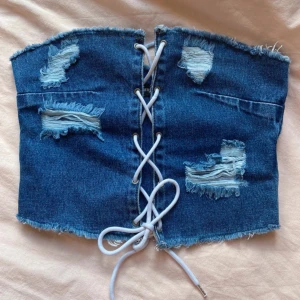 Jeans tubtopp med snörning  - Säljer en blå jeans tubtopp från PrettyLittleThing med snygga slitningar och råa kanter. Toppen har snörning framtill och är helt ärmlös. Perfekt för dig som vill sticka ut med en cool denimlook. Storleken är 38 men skulle säga att det är en S/XS och för någon med liten byst. 