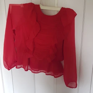 Röd volangblus från Lindex Kids - Superfin röd blus från Lindex Kids med långa ärmar och volangdetaljer framtill och vid midjan. Blusen är gjord i ett lätt och transparent tyg som ger en luftig känsla. Perfekt för dig som vill sticka ut med en färgstark och lekfull stil.