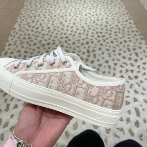 Beige och vita sneakers från Dior - Snygga sneakers från Dior i beige och vitt med ikoniskt mönster och broderad logga. Skorna har vit snörning, platt sula och rund tå. Perfekta för dig som vill ha en exklusiv och trendig look.