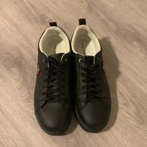 Svarta sneakers från Pier One - Svarta sneakers från Pier One med snygg broderad detalj i rött, blått och guld på sidan. Skorna har svarta snören, guldfärgade öljetter och vit insida. Röd rand på hälen ger en cool kontrast. Perfekta för dig som gillar stilrena men ändå unika skor.