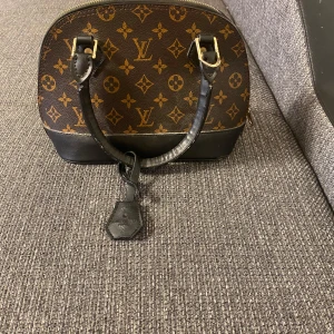 Louis Vuitton Monogram handväska - Säljer en klassisk Louis Vuitton handväska i brunt med ikoniskt monogrammönster och svarta skinnhandtag. Väskan har guldfärgade metalldetaljer och dragkedja upptill. Perfekt storlek för det viktigaste och en riktig statement piece.