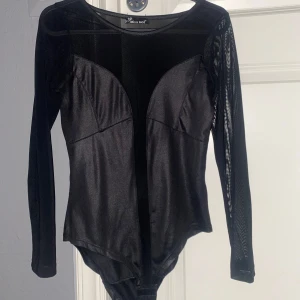 Svart långärmad bodysuit Miss Moi - Snygg svart bodysuit från Miss Moi med långärmade meshdetaljer och djup urringning. Toppen har en tight passform och är tillverkad i glansigt syntetmaterial som ger en cool look. Perfekt för att sticka ut med en edgy vibe.