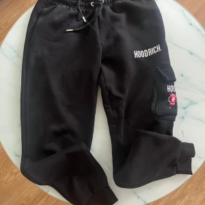 Svarta mjukisbyxor från Hoodrich - Svarta mjukisbyxor från Hoodrich med broderad logga på benet och praktisk ficka på sidan. Byxorna har resår och snörning i midjan samt muddar vid bensluten. Perfekta för en chill och avslappnad stil.