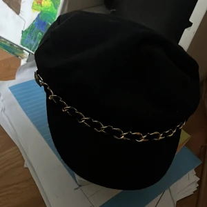 Svart vegamössa med guldkedja från Gina Tricot - Stilren svart vegamössa från Gina Tricot med snygg guldfärgad kedjedetalj framtill. Klassisk form med mjuk skärm och rundad krona. Tillverkad i polyester för en lätt och bekväm känsla. Perfekt accessoar för att lyfta din outfit. Superbra skick förutom lite djurhår på