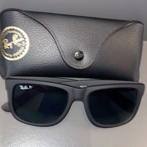 Svarta Ray-Ban solglasögon Wayfarer - Snygga svarta Ray-Ban Wayfarer solglasögon med mattsvart båge och mörka glas. Klassisk fyrkantig form och Ray-Ban-logga på linsen. Kommer med original svart fodral och putsduk. Perfekta för att lyfta din stil i solen.