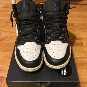 Nike Air Jordan 1 Mid White/ racer blue- black  - Nike Air Jordan 1 Mid sneakers i svart, blått och vitt skinn. Klassisk siluett med blå swoosh, svarta snören och perforerad tå. Ikonisk Jumpman-logga på plösen och Air Jordan-märke på sidan. Perfekta för dig som vill sticka ut med en tidlös streetstyle.