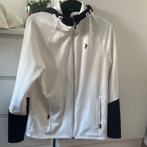 Vit vindjacka Peak Performance S - Snygg vit vindjacka från Peak Performance med svarta detaljer på ärmar och huva. Jackan har hel dragkedja, två fickor med dragkedja och en sportig look. Perfekt för dig som gillar stilren design och funktionella plagg.