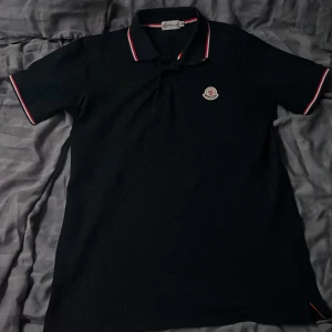 Moncler pike - Säljer en stilren svart pikétröja från Moncler med röd och vit randig detalj på krage och ärmslut. Tröjan har en klassisk passform och Moncler-logga på bröstet. Perfekt för en avslappnad men snygg look.