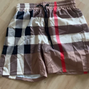 Rutig Burberry badshorts med snörning - Snygga badshorts från Burberry med klassiskt rutigt mönster i beige, svart, vitt och rött. Shortsen har elastisk midja med svart snörning och innerbyxa i vitt mesh. Perfekta för sommaren och ger en lyxig vibe på stranden.