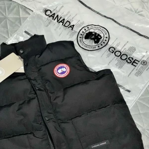 Svart dunväst från Canada Goose - Säljer en svart dunväst från Canada Goose med klassisk logga på bröstet. Västen har hög krage, två stora fickor framtill och stängs med både dragkedja och knappar. Perfekt för lager-på-lager och riktigt snygg till streetwear.