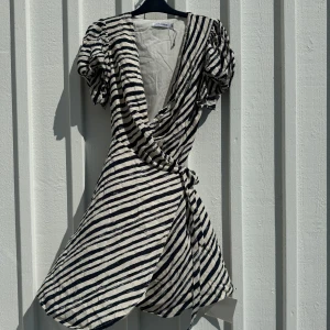 Randig omlottklänning från Adoore - Kort omlottklänning från Adoore med svartvita diagonala ränder. Klänningen har puffiga korta ärmar och knyt i midjan. Aldrig använd. Strl 42