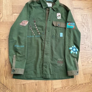 Grön overshirt från Scotch & Soda - Snygg grön overshirt från Scotch & Soda med broderade detaljer som palmer, blommor och texten 'Aloha Spirit' och 'No worries'. Jackan har patch på ärmen, bröstficka och coola kontrastdetaljer vid manschetterna. Perfekt för dig som gillar unika plagg med tropisk vibe. Jackan är införskaffad från en Scotch & Soda butik i Frankrike. Nypris: 400 euro, alltså ca 4000 kr. Jackan finns inte längre och är extremt snygg. Hör av dig vid minsta frågetecknet! 