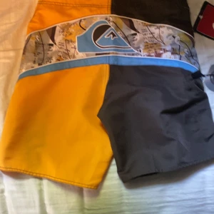Quiksilver Kelly Slater badshorts - Snygga Quiksilver badshorts från Kelly Slater-kollektionen. De är tvåfärgade i gult och brunt med coolt grafiskt mönster och blå detaljer. Klassisk boardshort-modell med snörning i midjan och sidoficka med dragkedja. Perfekta för stranden eller poolen!    Helt nya, men har redan 2 andra så denna behövde bli såld STORLEK 2,8