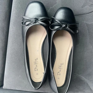 Svarta ballerinaskor från Duffy - Klassiska svarta ballerinaskor från Duffy med rund tå och dekorativ rosett framtill. Skorna har en platt sula och är tillverkade i syntetmaterial med en stilren look som passar till många outfits. Helt nya