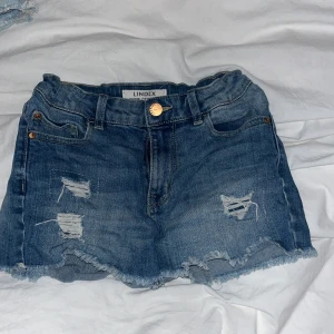 Blå jeanskjol med slitningar  - Snygga blå jeanskjol från Lindex med råa kanter och slitna detaljer framtill. Klassisk femficksmodell med knapp och dragkedja. Perfekta för varma dagar när du vill ha en avslappnad och trendig look.