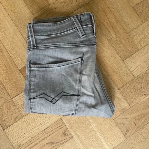 Replay grå jeansbyxor slim fit - Snygga grå jeans från Replay med klassisk femficksdesign och svarta detaljer på bakfickan. Jeansen har en slim passform och raka ben, tillverkade i mjukt bomullsmaterial med lite stretch för extra komfort. Perfekta för en clean och stilren look.