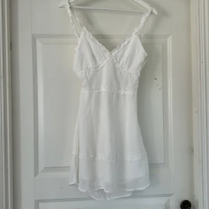 Vit kort klänning från H&M Divided - Jag säljer en Supersöt vit kort klänning från H&M Divided. Klänningen har smala axelband, volangdetaljer och en snygg knytning i ryggen💗