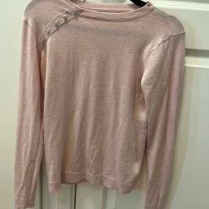 Ljusrosa tröja med pärlknappar Mohito - Supersöt ljusrosa långärmad tröja från Mohito med dekorativa pärlknappar på ena axeln. Tröjan är tunnstickad och har en klassisk, rak passform som är lätt att styla till olika looks. Perfekt för dig som gillar feminina detaljer och mjuka färger. XXS men sitter lika bra på mig som har M