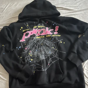 Svart hoodie SP5DER med spindelnät - Svart hoodie från SP5DER i 100% bomull med stor spindelnätsgrafik och texten 'pink!' i rosa framtill. Hoodien har huva, känguruficka och färgglada stjärnor och detaljer. På ryggen finns en gul stjärnlogga. Perfekt statement-plagg för dig som gillar streetwear.