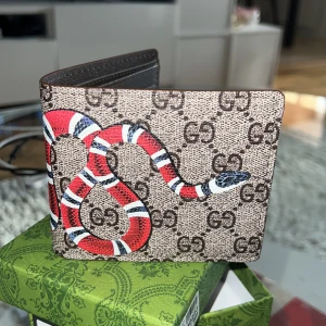 Ny plånbok med röd orm - Snygg plånbok med klassiskt GG-mönster och en cool röd, vit och svart orm på framsidan. Plånboken är i brunt skinn och har flera kortfack samt fönster för ID. 