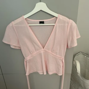 Ljusrosa topp  - Supersöt ljusrosa topp från Gina Tricot med v-ringning och korta, lite fladdriga ärmar. Toppen har knytband i midjan och är croppad för en trendig look. Tillverkad i mjuk viskosblandning som känns lätt och skön mot huden.