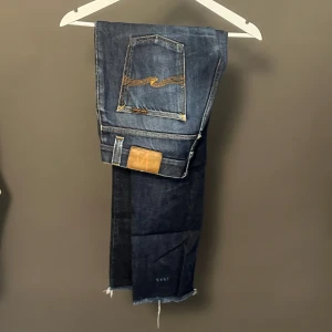 Nudie Jeans Grim Tim Dry Dirt 32/34 - Säljer ett par Nudie Jeans Grim Tim Dry Dirt i mörkblå tvätt med snygga slitningar och klassiska orange sömmar. Jeansen har raka ben, fem fickor och är tillverkade i ekologisk bomull. Perfekt för dig som gillar en clean men ändå cool look.