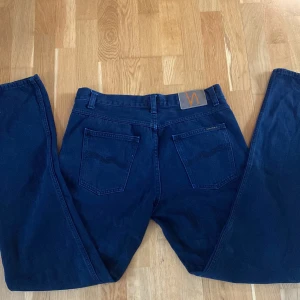 Mörkblå jeans från Nudie Jeans - Säljer ett par Skit Feta mörkblå jeans från Nudie Jeans med klassisk femficksdesign och snygga detaljer på bakfickorna. Jeansen har rak passform och är tillverkade i bomull. Perfekta för dig som gillar stilrena och tidlösa jeans.