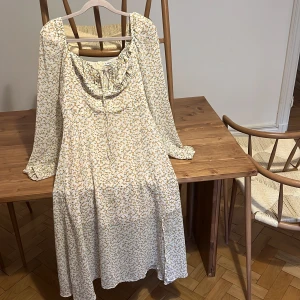 Blommig långklänning från Adoore - Supersöt långklänning från Adoore i ljusbeige med små gula blommor. Klänningen har långa ärmar, söt knytning vid urringningen, slits i sidan och dragkedja bak. Perfekt flowy passform och romantisk vibe.