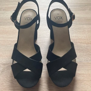 Svarta sandaler med klack från VOX - Snygga svarta sandaler från VOX med breda korsade remmar och justerbart spänne i guldfärg. Skorna har en stabil hög klack och öppen tå. Tillverkade i veganskt material med mockakänsla, perfekta för att lyfta din outfit. Köpt för 400kr och endast använt en gång. Storlek 39.