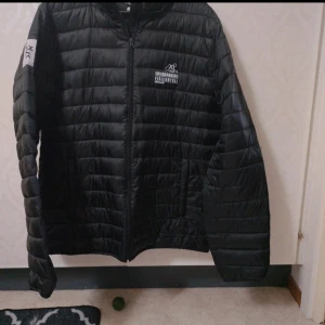 Svart dunjacka EriksGårdens Fjällhotell - Svart dunjacka med quiltad design och dragkedja framtill. Jackan har tryck med Eriksgårdens Fjällhotell Funäsdalen på bröstet och en vit patch med texten 'eriks kök & bar' på ärmen. Perfekt för kalla dagar och riktigt skön passform.