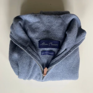 Blå kashmirtröja från Alan Flusser - Mjuk och lyxig ljusblå tröja i 100% kashmir från Alan Flusser. Tröjan har half zip-dragkedja med läderdetalj, ribbade muddar och lång ärm. Perfekt för dig som gillar stilren och bekväm look. Säljer för 700kr nypris 2200kr, storleken är L men passar mer som en M
