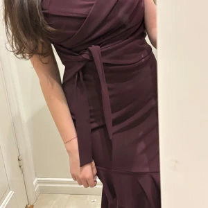 Vinröd omlottklänning med volang - Säljer en vinröd omlottklänning med knyt i midjan och volangdetalj nertill. Klänningen har en figurnära passform och är ärmlös, perfekt för dig som vill ha en elegant och stilren look. Klänningen passar till alla evenemang, och sitter som en smäck. Passar till dig som har storlek mellan  34 och 36🫶🏼