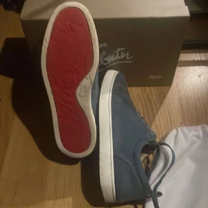 Blå sneakers från Christian Louboutin - Säljer ett par blå sneakers från Christian Louboutin med klassisk röd sula och vita detaljer. Skorna har snörning och rund tå, och är tillverkade i mocka. Perfekta för dig som vill sticka ut med stil.