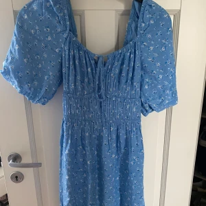 Blå blommig klänning från BikBok - Helt oanvänd! Charmig blå klänning från BikBok med ett sött blommönster. Klänningen har korta puffärmar och en knytning framtill. Perfekt för en somrig look! 