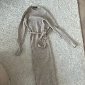 Beige ribbad klänning med bälte - Snygg beige ribbad klänning med lång ärm och matchande bälte i midjan. Perfekt för en stilren look. Klänningen har en klassisk rund hals och är gjord i ett mjukt material som ger en bekväm passform.