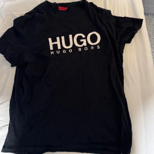 Svart t-shirt från Hugo Boss - Snygg svart t-shirt från Hugo Boss med stort tryck på framsidan. Perfekt för en stilren och avslappnad look. Passar bra till jeans eller shorts.