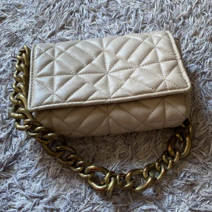 Beige quiltad axelväska med kedja - Snygg beige axelväska med quiltat mönster och en rejäl guldfärgad kedja som axelrem. Väskan har ett praktiskt magnetlås och ett rymligt inre fack. Perfekt för att ge din outfit en stilfull touch. Kan gå ner i pris vid snabb affär💖