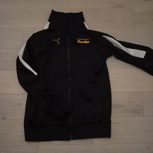 Svart Porsche Turbo x Puma track jacket - Svart track jacket från Puma i samarbete med Porsche Turbo. Jackan har vita ränder längs ärmarna, gul Porsche Turbo-logga på bröstet, Puma-logga och en gul bilsilhuett på ärmen. Hög krage och dragkedja framtill.